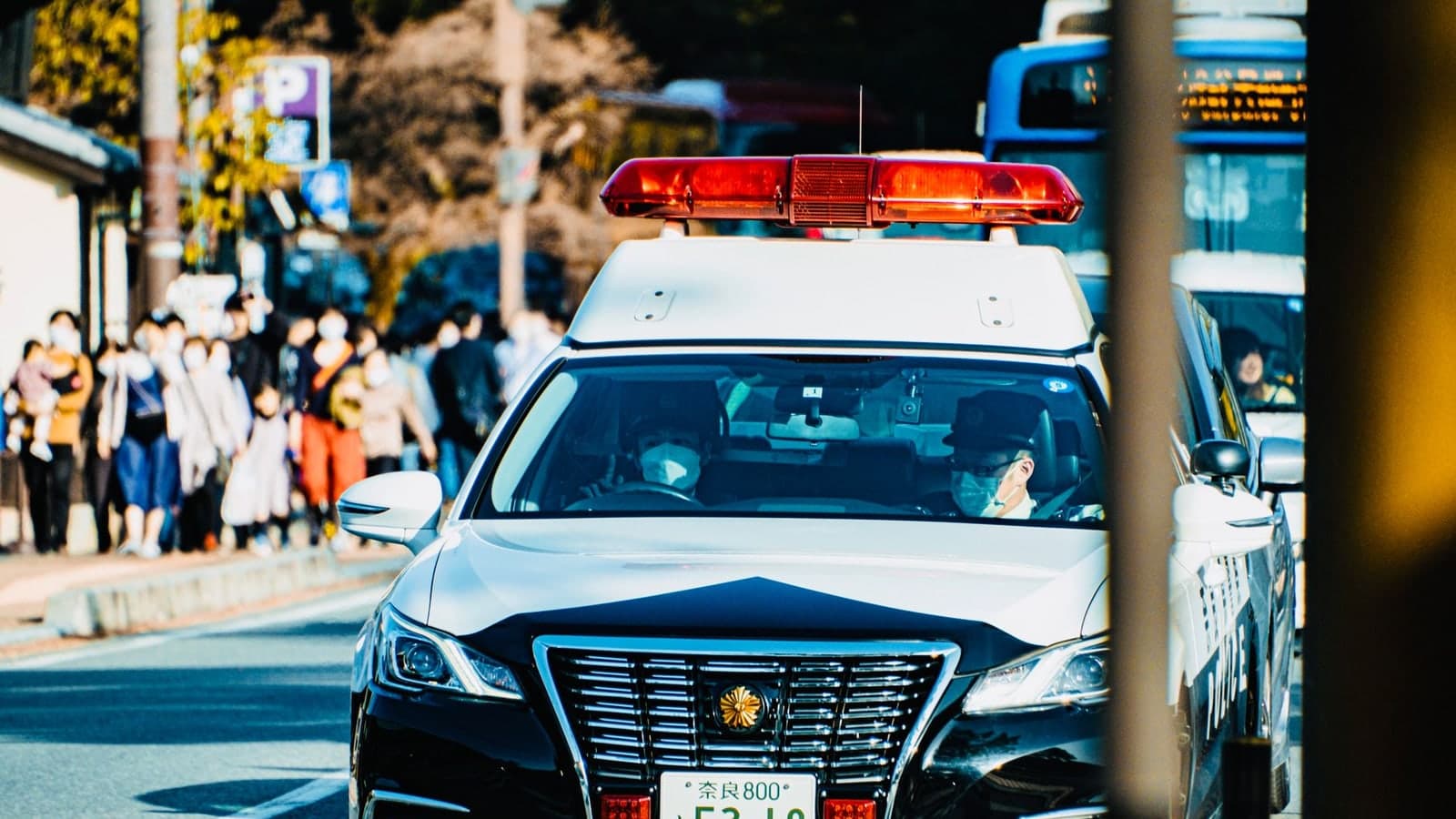 A police vehicle with flashing lights is parked on a street people standing nearby, and another is partially visible to the right — illustrative photograph