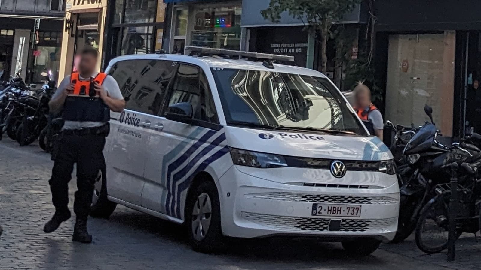 Police officers stand beside a white volkswagen van parked on a cobblestone street with motorcycles and bicycles nearby — illustrative photograph