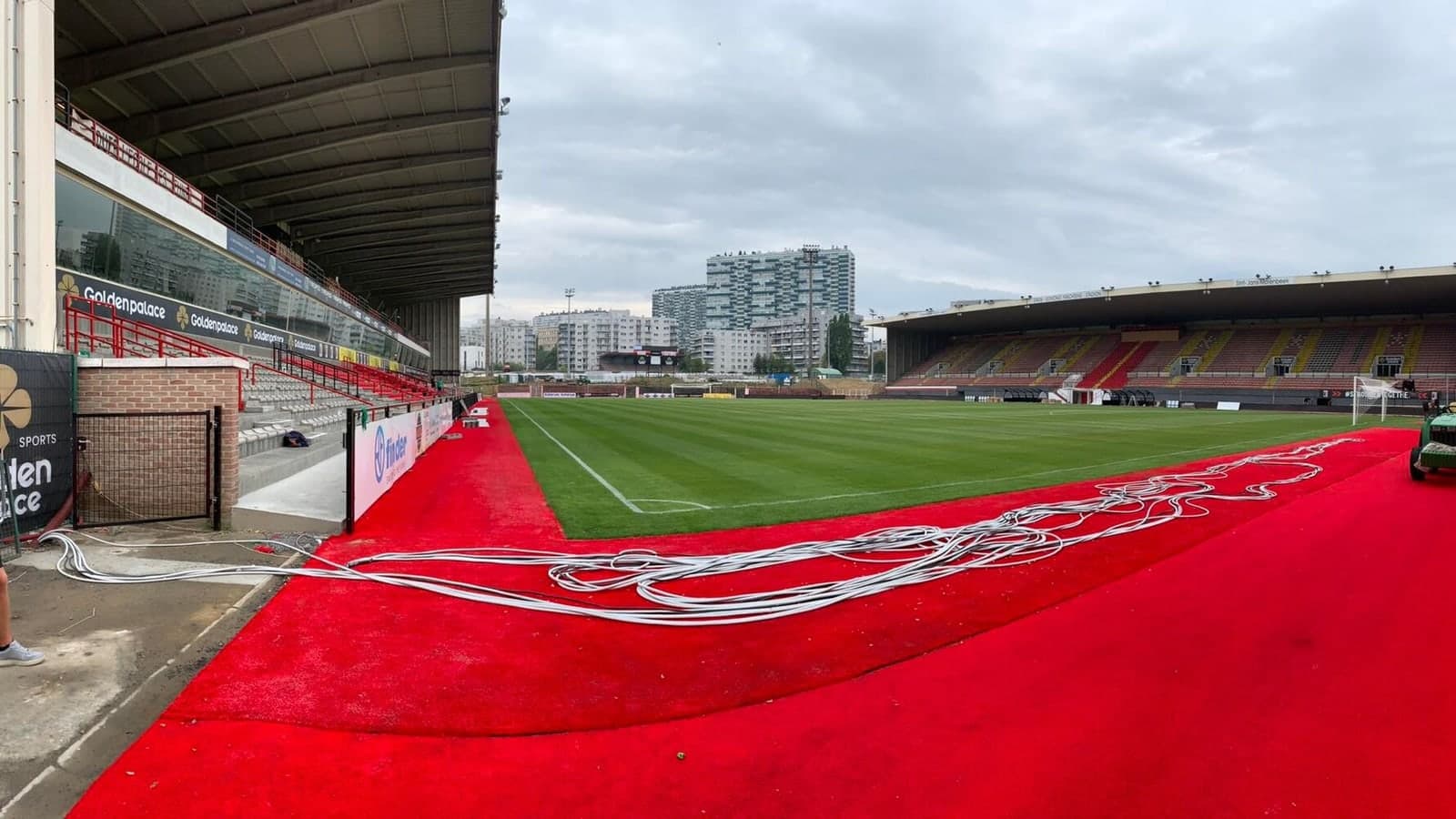 An empty football stadium with a red carpet laid out on the sidelines and coiled white cables nearby, under an overcast sky — illustrative photograph