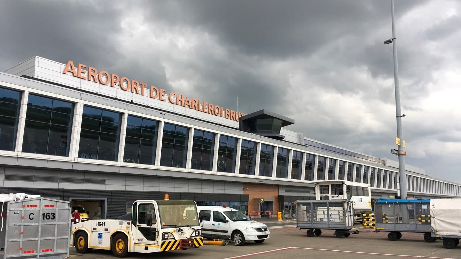 A modern airport terminal under a cloudy sky with ground support equipment parked in front — illustrative photograph