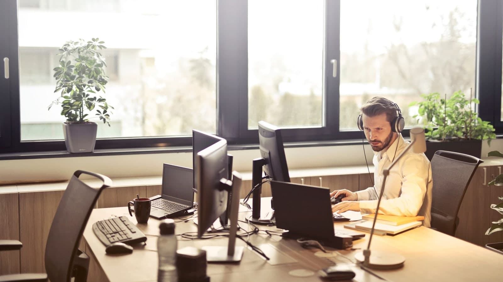 A businessman sits at a desk using multiple computers and a headset in a well-lit modern office — illustrative photograph