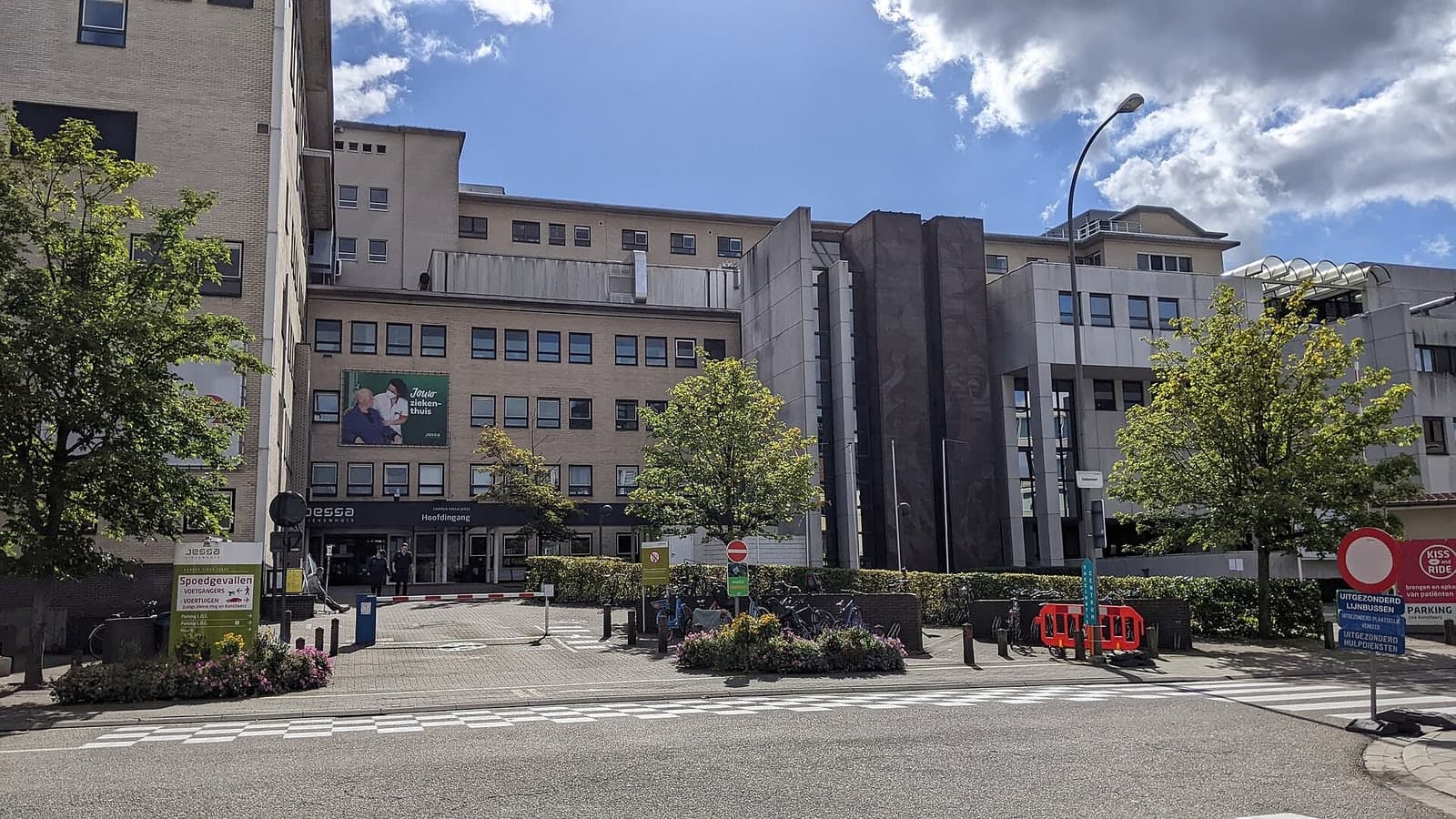 A modern hospital complex with multiple buildings under a partly cloudy sky, surrounded by trees and paved areas — illustrative photograph