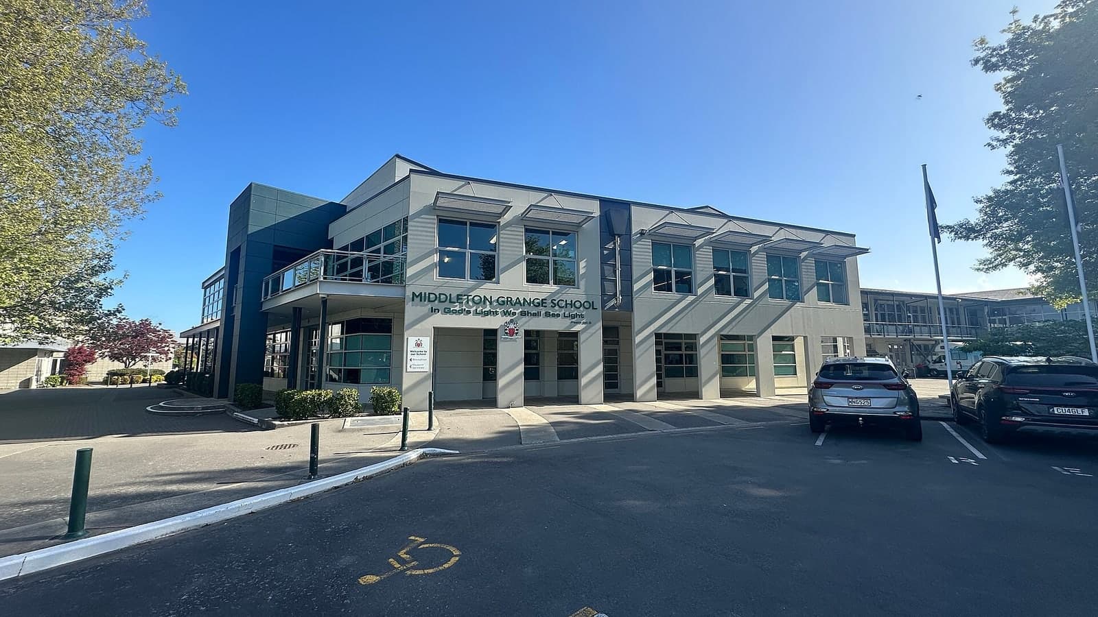 A modern school building under clear blue skies with parked cars and flagpoles in front — illustrative photograph