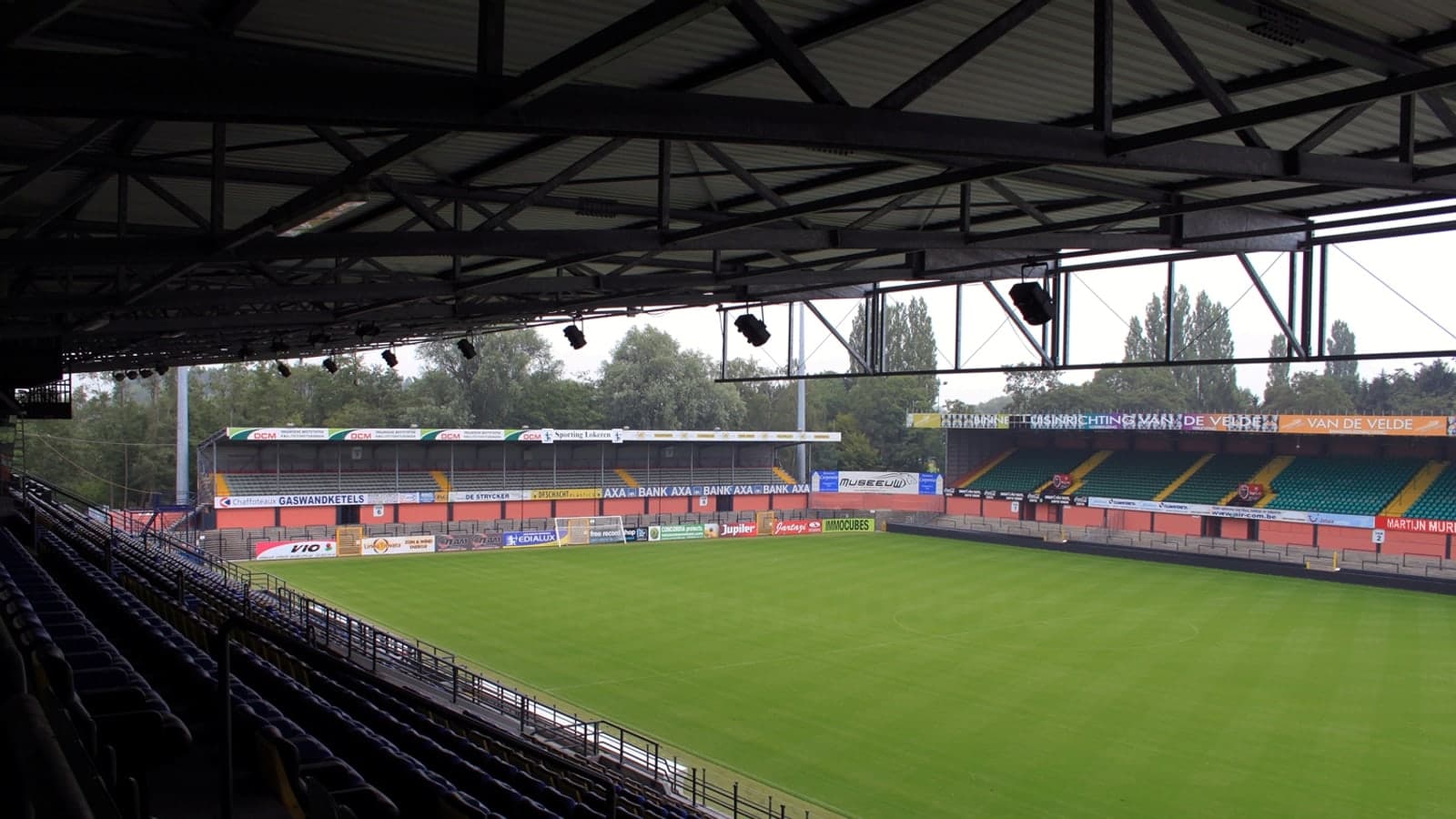An empty football stadium with green grass and tiered seating under a metal roof, surrounded by trees — illustrative photograph