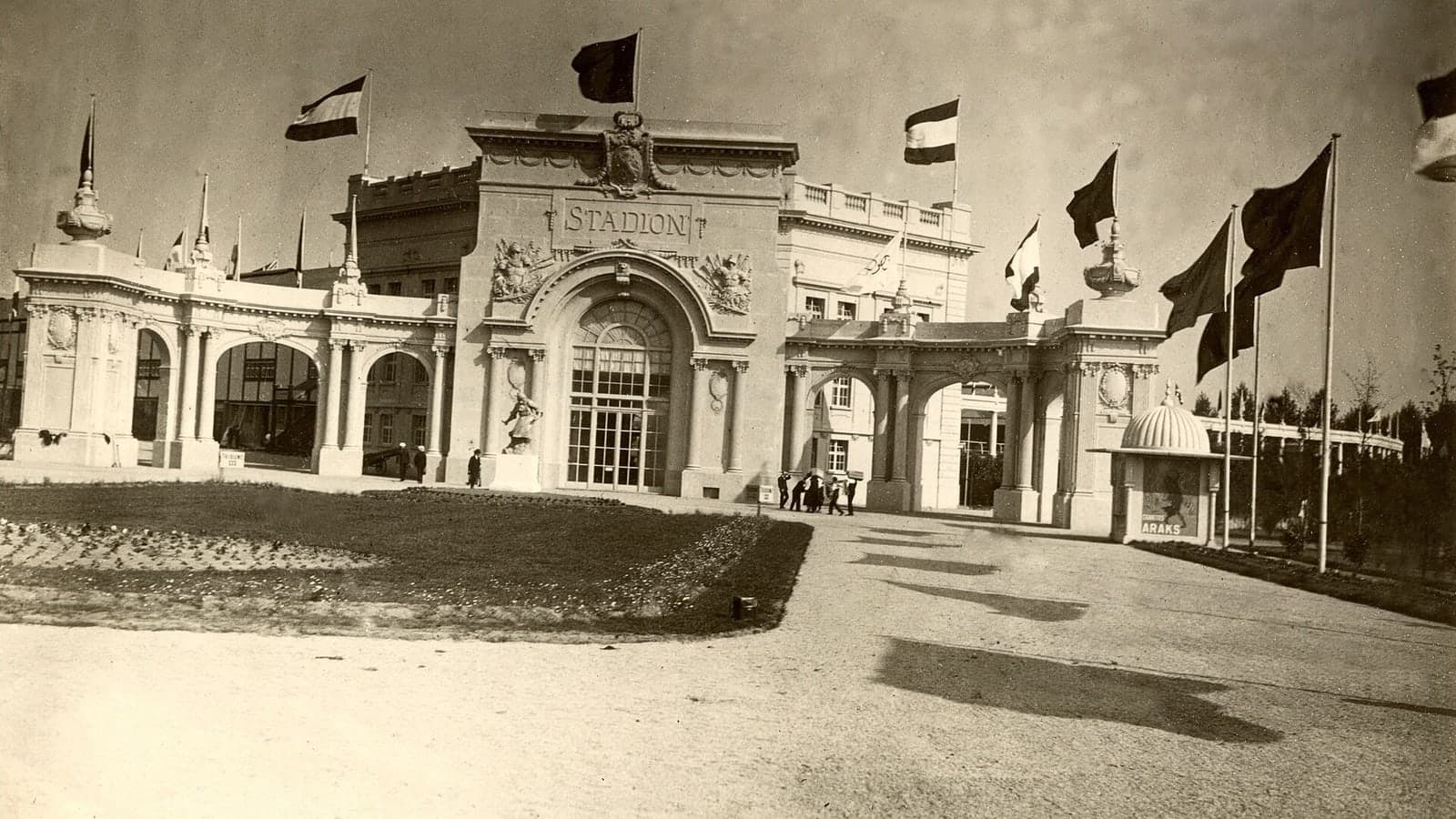 A grand, ornate stadium entrance with multiple flags flying and people walking nearby — illustrative photograph