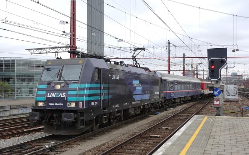 An electric locomotive pulling passenger cars stops at a railway station with overhead wires and signaling equipment visible — illustrative photograph