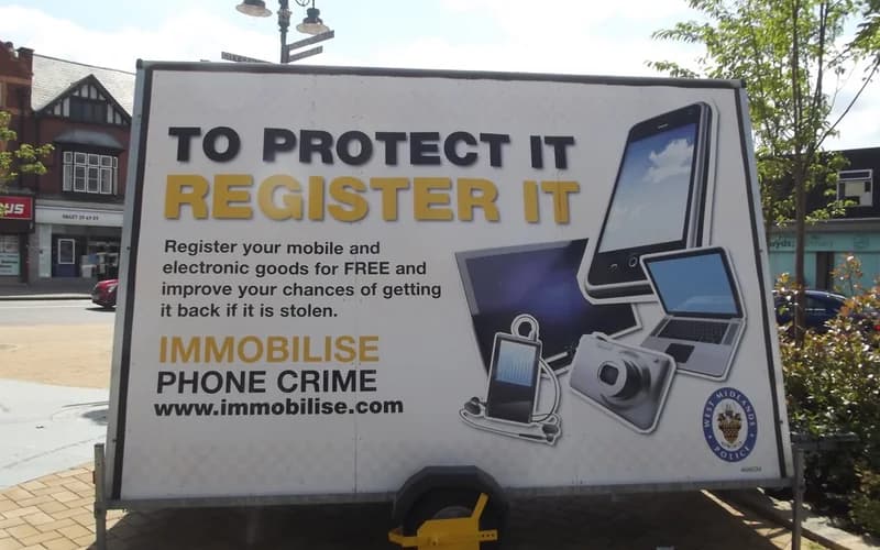 A public billboard promoting mobile phone registration to prevent theft, displayed outdoors near buildings and trees — illustrative photograph